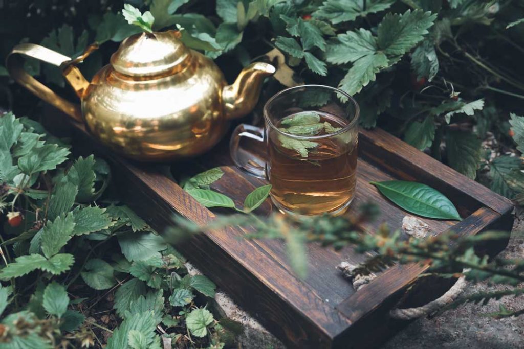 Close-up view of a wooden platter box on green leaves, showcasing a clear tea cup filled with Moroccan Mint Tea. Mint and green tea leaves are scattered on the platter, with a golden teapot placed next to the cup.